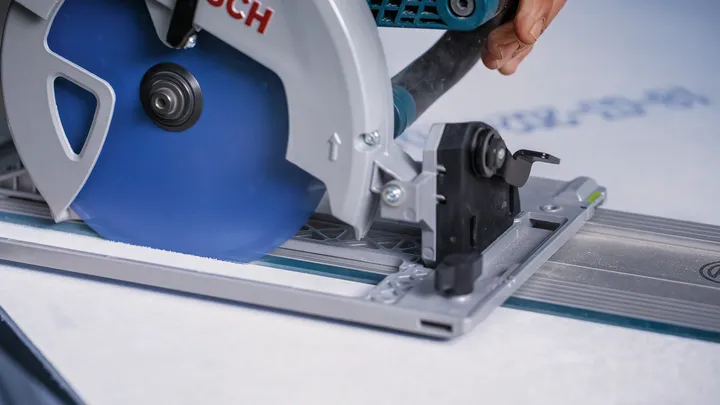 Una persona che indossa un equipaggiamento di sicurezza guida una sega circolare mentre taglia il cartongesso lungo una guida metallica.