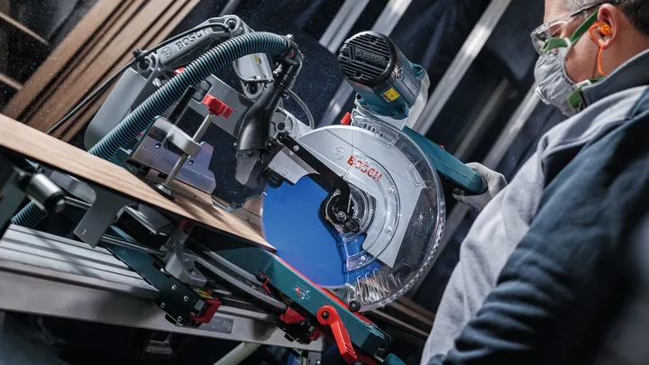 Una persona che indossa dispositivi di sicurezza utilizza una sega circolare per tagliare una tavola di legno.