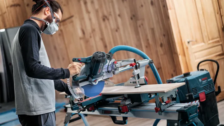 Una persona che indossa dispositivi di sicurezza taglia il legno con una sega troncatrice in un'officina.