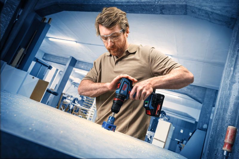 Una persona che indossa un equipaggiamento di sicurezza fora una superficie di lavoro con un trapano a batteria.