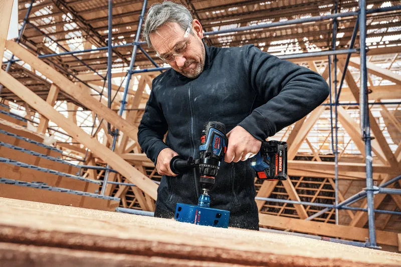 Una persona che indossa un equipaggiamento di sicurezza pratica un grande foro nel legno con un trapano a batteria.