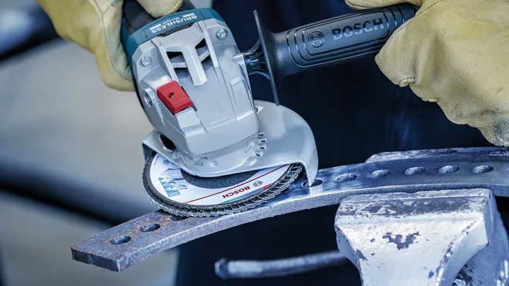 Una persona che indossa un equipaggiamento di sicurezza macina una barra di metallo con un utensile elettrico.