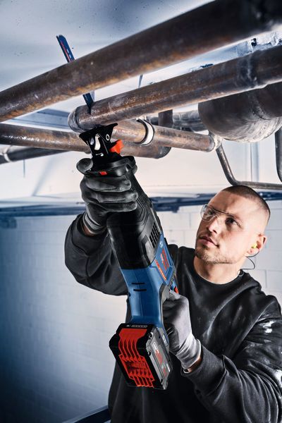 Una persona che indossa dispositivi di sicurezza taglia tubi metallici sospesi in alto con un seghetto alternativo a batteria.