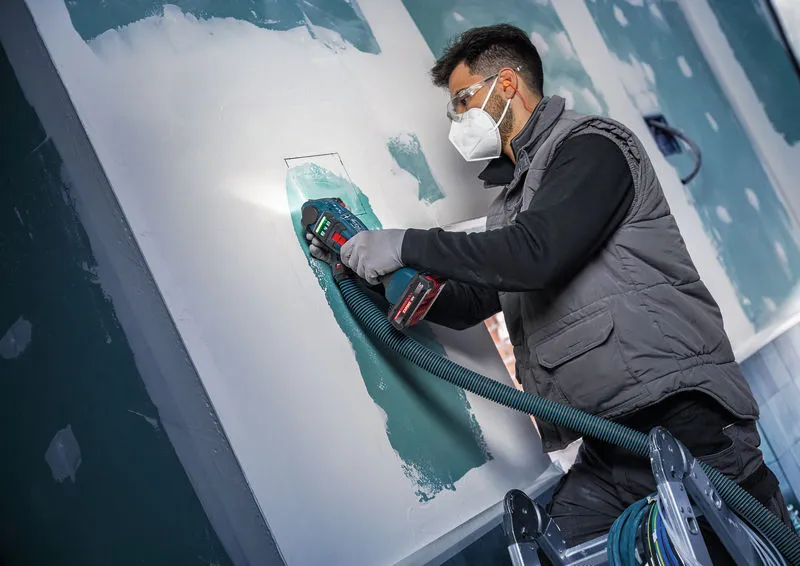 Una persona che indossa un equipaggiamento di sicurezza utilizza un multi-cutter senza fili per tagliare il cartongesso.