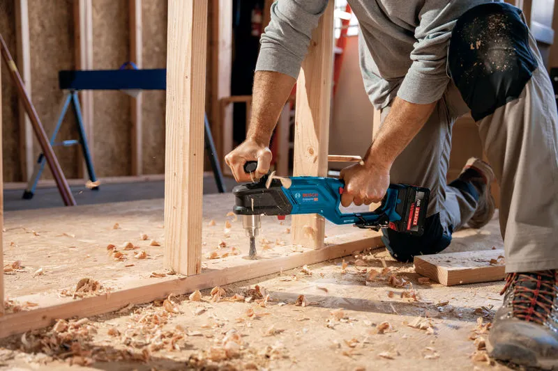 Una persona che indossa dispositivi di sicurezza fora un telaio di legno con un trapano angolare a batteria.