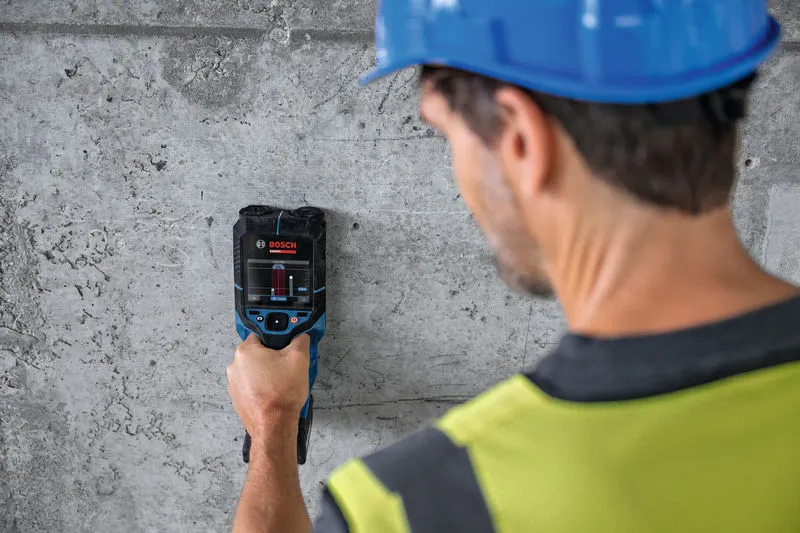 Una persona che indossa un equipaggiamento di sicurezza esamina un muro di cemento con un dispositivo di rilevamento.