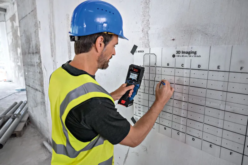 Una persona che indossa un equipaggiamento di sicurezza utilizza un rilevatore per scansionare e contrassegnare una griglia su un muro.