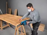 Una persona che indossa dispositivi di sicurezza leviga una porta di legno con una levigatrice a nastro.