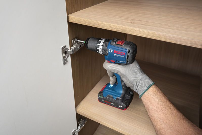 Una persona che indossa dispositivi di sicurezza utilizza un trapano a percussione a batteria per installare la cerniera di un mobile.