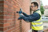Una persona che indossa un equipaggiamento di sicurezza fora un muro di mattoni utilizzando un trapano a percussione a batteria.