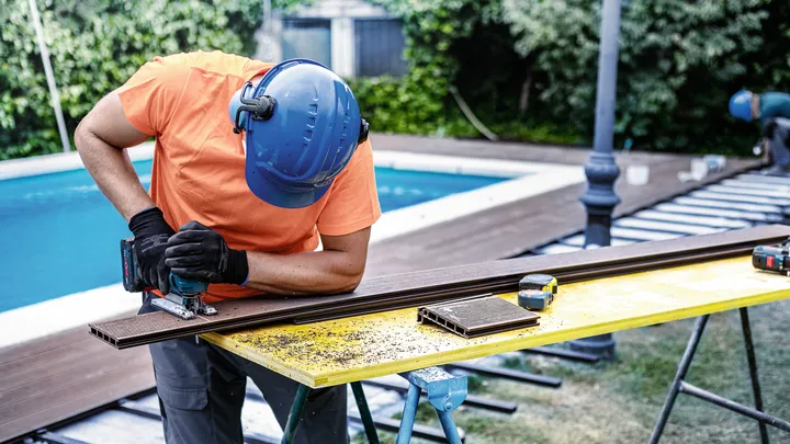 Una persona che indossa dispositivi di sicurezza utilizza un seghetto alternativo per tagliare le assi del terrazzo all'aperto.
