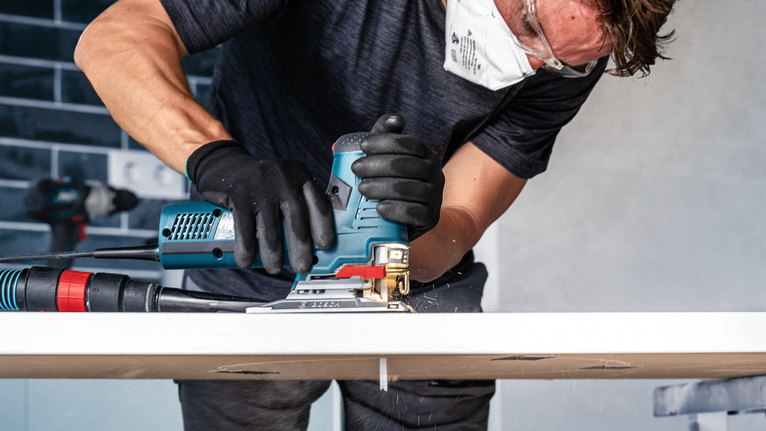 Una persona che indossa un equipaggiamento di sicurezza usa un seghetto alternativo per tagliare una tavola di legno.