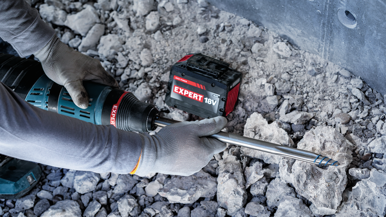 Una persona che indossa dispositivi di sicurezza aziona un martello perforatore a batteria su un terreno roccioso.