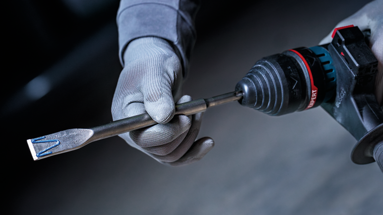 La persona che indossa l'equipaggiamento di sicurezza tiene in mano un trapano a percussione rotante dotato di punta a scalpello.