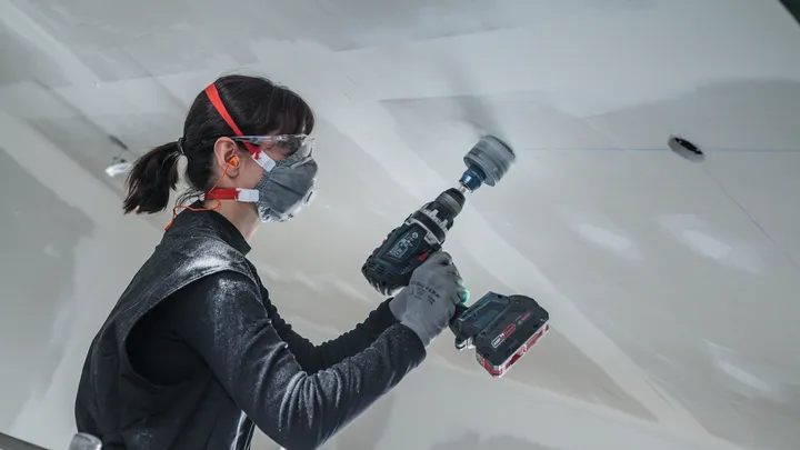 Una persona che indossa dispositivi di sicurezza utilizza un trapano a batteria per tagliare un soffitto.
