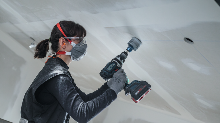 Una persona che indossa dispositivi di sicurezza utilizza un trapano a batteria per tagliare un soffitto.
