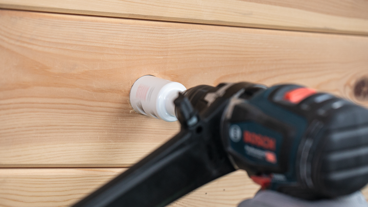 Una persona che indossa un equipaggiamento di sicurezza pratica un foro nel legno con un utensile elettrico.