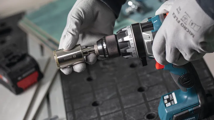 Una persona che indossa un equipaggiamento di sicurezza collega una sega a tazza a un trapano a batteria.