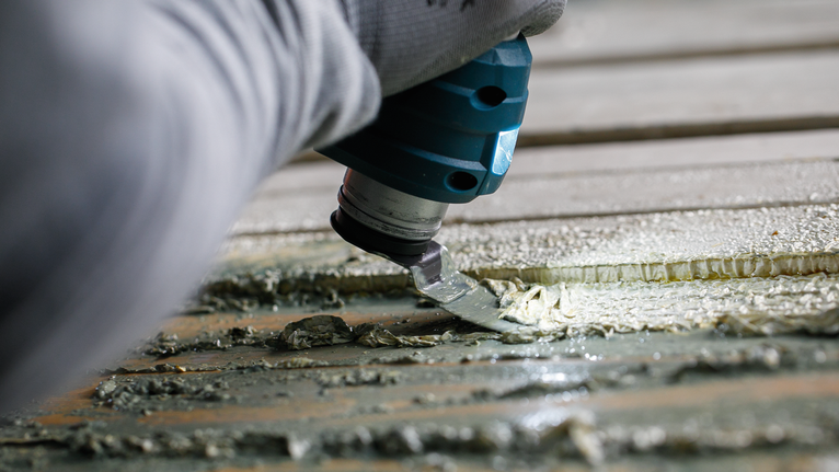 Una persona che indossa un equipaggiamento di sicurezza utilizza un utensile multiuso per raschiare l'adesivo da una superficie.