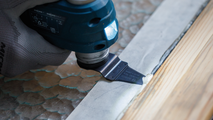 Una persona che indossa un equipaggiamento di sicurezza utilizza un utensile multiuso per tagliare lungo un bordo di legno.