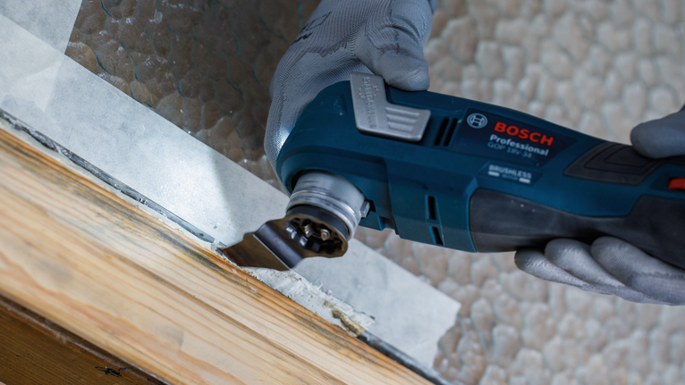 Una persona che indossa dispositivi di sicurezza leviga il telaio di una finestra in legno utilizzando un utensile elettrico.