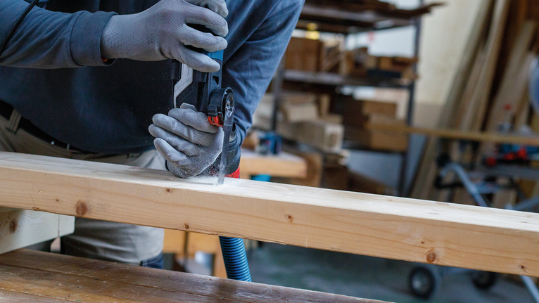 Una persona che indossa un equipaggiamento di sicurezza taglia una trave di legno con un utensile elettrico.