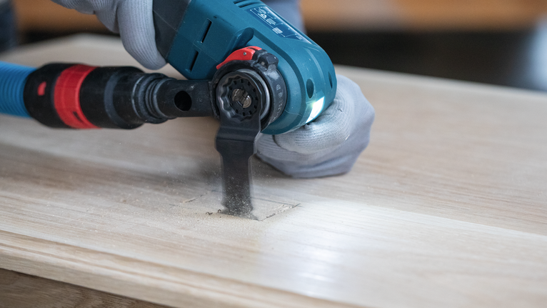 Una persona che indossa un equipaggiamento di sicurezza utilizza un elettroutensile per praticare un foro rettangolare nel legno.