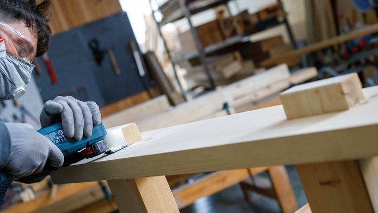 Una persona che indossa l'equipaggiamento di sicurezza carteggia una tavola di legno con un elettroutensile.