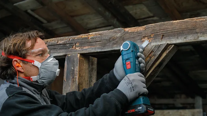 Persona che indossa dispositivi di sicurezza mentre utilizza un utensile senza fili per levigare una trave di legno.