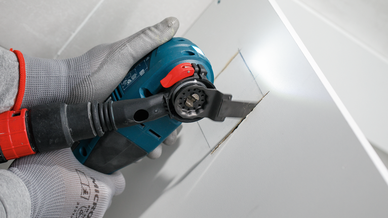 Una persona che indossa un equipaggiamento di sicurezza taglia un'apertura rettangolare nel muro a secco con un utensile elettrico.