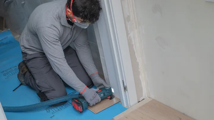 Una persona che indossa dispositivi di sicurezza sta rifinendo il pavimento con un elettroutensile vicino allo stipite di una porta.