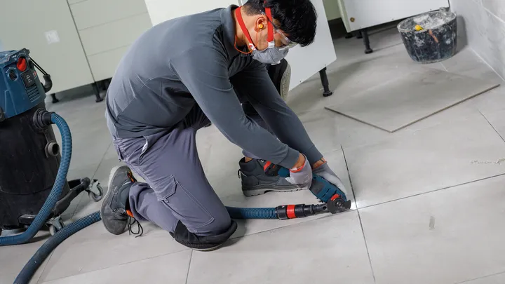 Una persona che indossa dispositivi di sicurezza taglia le piastrelle del pavimento con un elettroutensile collegato a un aspirapolvere.
