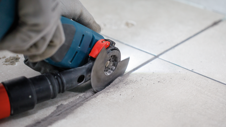 Persona che indossa dispositivi di sicurezza e utilizza un elettroutensile per tagliare le piastrelle lungo una linea di fuga.
