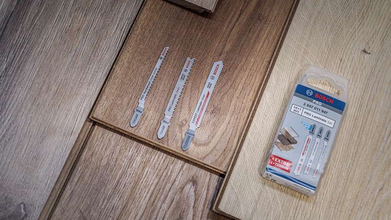 Tre lame per seghetto alternativo e il loro imballaggio appoggiati su un pavimento in laminato di legno.