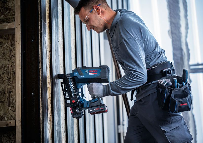 Una persona che indossa un equipaggiamento di sicurezza utilizza una chiodatrice per legno senza fili su un telaio verticale.