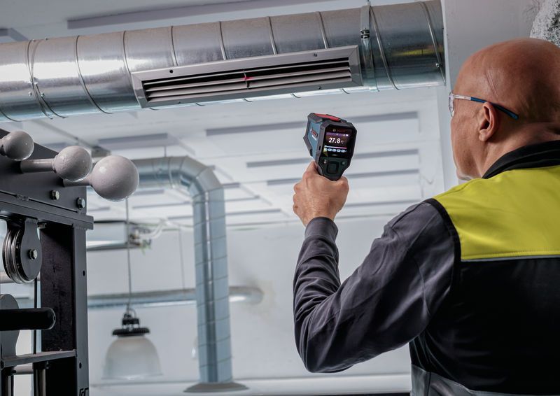 Una persona che indossa dispositivi di protezione individuale utilizza un rilevatore di temperatura per controllare la temperatura in prossimità di una bocchetta di ventilazione.