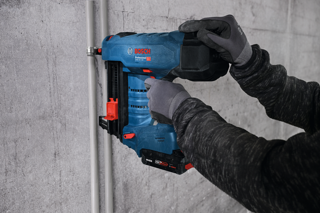 Una persona che indossa un equipaggiamento di sicurezza utilizza una chiodatrice a batteria per fissare un condotto a un muro di cemento.