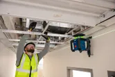 Una persona che indossa dispositivi di sicurezza installa una struttura per soffitto utilizzando uno strumento di livellamento laser.