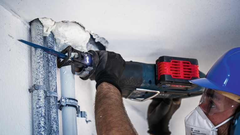 Una persona che indossa un equipaggiamento di sicurezza taglia un tubo del soffitto utilizzando un seghetto alternativo.