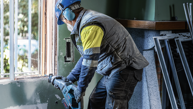 Una persona che indossa un equipaggiamento di sicurezza utilizza un elettroutensile per tagliare il telaio di una finestra.