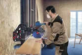 Una persona che indossa dispositivi di protezione individuale utilizza una sega a troncare per tagliare una tavola di legno.