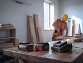 Batterie e caricabatterie per utensili elettrici su un banco da lavoro, con una persona che indossa dispositivi di protezione individuale intenta ad assemblare del legno.