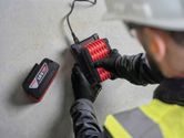 Una persona che indossa dispositivi di protezione individuale collega una batteria a un caricabatterie su una superficie di cemento.