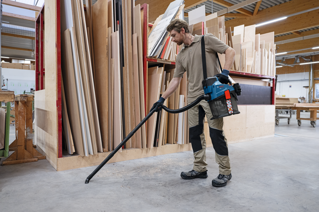 Una persona con indosso dispositivi di protezione individuale utilizza un aspirapolvere portatile per pulire il pavimento di un'officina di falegnameria.