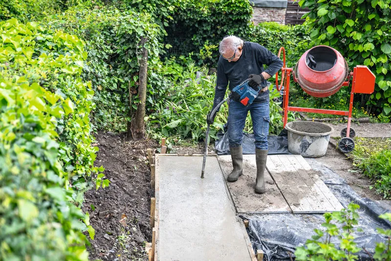 Una persona che indossa dispositivi di protezione individuale utilizza un utensile elettrico per compattare il calcestruzzo su un vialetto da giardino.