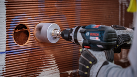 Una persona che indossa dispositivi di protezione individuale utilizza una sega a tazza per praticare un foro in una parete piastrellata.