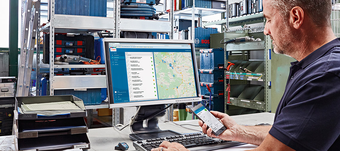 Mann am Computer nutzt Smartphone, Kartendaten auf Bildschirm, Werkstatt im Hintergrund.