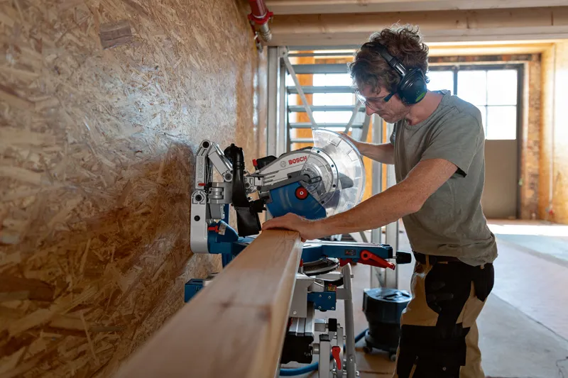 Una persona que usa equipo de seguridad corta una viga de madera utilizando una sierra ingletadora inalámbrica.