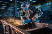 Una persona que usa equipo de seguridad muele vigas de metal con una amoladora angular, creando chispas.