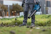 Una persona que lleva equipo de seguridad utiliza un soplador de hojas inalámbrico sobre el césped.